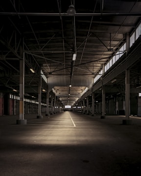 A large, empty industrial warehouse with high ceilings, exposed steel beams, and a symmetrical layout. The interior is dimly lit, with patches of light creating an interplay of shadows on the floor. Rows of support columns stretch into the distance, and narrow windows line the upper walls, allowing minimal natural light.