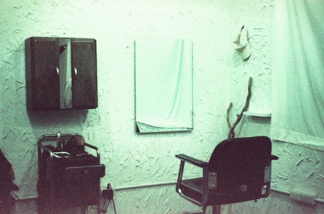 A dark salon chair and a small trolley beside a wall mirror and a wall cabinet, in a room with textured green-tinted walls