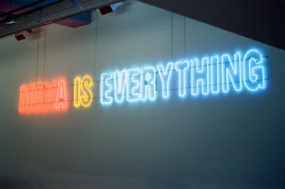 Neon lights display the phrase 'MEDIA IS EVERYTHING' in a sequence of red, yellow, and blue colors against a plain wall. The glow of the neon creates a vibrant and striking visual, infusing the scene with a modern and edgy look.