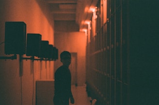A dimly lit hallway with a series of speakers on the left wall, illuminated by soft orange light. A silhouette of a person is visible in the center, creating a mysterious atmosphere. The perspective of the image draws attention to the depth of the hallway.