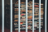 A large stack of books is secured with metal chains, placed behind metal bars. The books appear to be old and are arranged in an uneven pile, suggesting a feeling of confinement and restriction.