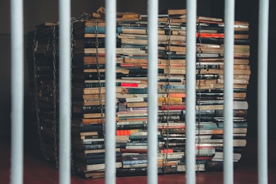 A large stack of books is secured with metal chains, placed behind metal bars. The books appear to be old and are arranged in an uneven pile, suggesting a feeling of confinement and restriction.