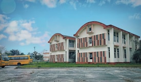 A two-story building with an architectural design featuring curved roofs and striped orange and white patterns. The building is surrounded by greenery and a concrete pathway in the foreground. Two yellow school buses are parked on the left side, with trees in the background under a partly cloudy blue sky.