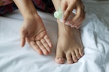 A close-up of a person's foot and their hands holding a small plastic bottle with a green cap. The person appears to be applying a liquid, possibly lotion or sanitizer, to their hand. The setting is on a white fabric surface, likely a bed or towel.