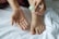 A close-up of a person's foot and their hands holding a small plastic bottle with a green cap. The person appears to be applying a liquid, possibly lotion or sanitizer, to their hand. The setting is on a white fabric surface, likely a bed or towel.