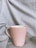 A light pink ceramic mug is placed on a soft, wrinkled, gray fabric background. The mug has a simple, smooth design with a rounded handle.