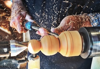 Hands operating a wood lathe, meticulously crafting and shaping a piece of wood amidst flying wood shavings.
