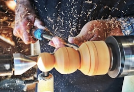 Hands operating a wood lathe, meticulously crafting and shaping a piece of wood amidst flying wood shavings.