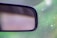 A close-up view of a car's rearview mirror with a blurred background. The mirror is positioned on the left side of the image, with a dotted pattern visible partially across the top section. The surrounding background features a combination of greens and soft focus light spots, giving a sense of looking out from inside a vehicle.
