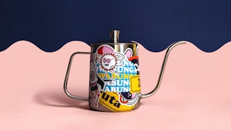 A metal teapot features colorful, whimsical stickers covering its surface. The handle and spout are long and curved. The background has a wavy design with a navy blue upper section and light pink lower portion, creating a playful contrast.