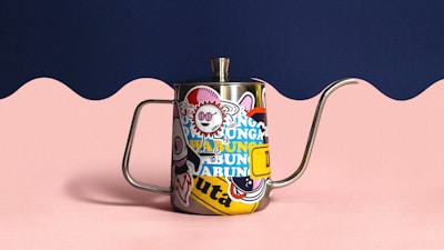 A metal teapot features colorful, whimsical stickers covering its surface. The handle and spout are long and curved. The background has a wavy design with a navy blue upper section and light pink lower portion, creating a playful contrast.