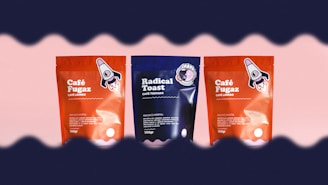 Three coffee packages are arranged side by side against a light pink background. The two outer packages are red with a rocket illustration, labeled 'Café Fugaz'. The center package is blue with a different design, labeled 'Radical Toast'. A wave-like dark border frames the top and bottom edges of the image.