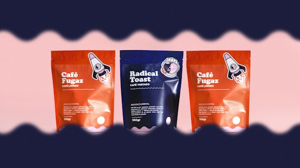 Three coffee packages are arranged side by side against a light pink background. The two outer packages are red with a rocket illustration, labeled 'Café Fugaz'. The center package is blue with a different design, labeled 'Radical Toast'. A wave-like dark border frames the top and bottom edges of the image.