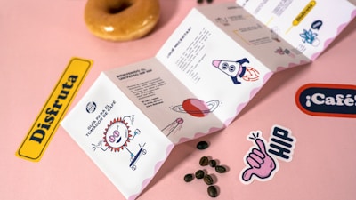 A playful and colorful scene featuring an illustrated pamphlet with various whimsical graphics, coffee beans scattered on a pink surface, and stickers with words and drawings. A glazed donut is positioned at the top.