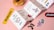 A playful and colorful scene featuring an illustrated pamphlet with various whimsical graphics, coffee beans scattered on a pink surface, and stickers with words and drawings. A glazed donut is positioned at the top.