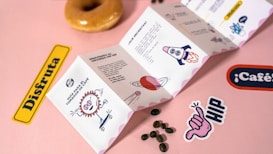 A playful and colorful scene featuring an illustrated pamphlet with various whimsical graphics, coffee beans scattered on a pink surface, and stickers with words and drawings. A glazed donut is positioned at the top.