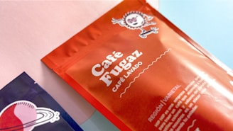 Two coffee bags are displayed on a surface. One bag is primarily orange with a design of a whimsical sun-like character and the text 'Café Fugaz'. The other bag, partially visible, is dark blue with a retro-style planet design. The background is a combination of pastel pink and blue colors.