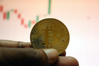 Hands holding a physical bitcoin coin with financial charts in the background.