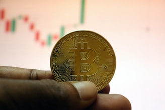 A hand holding a physical representation of a Bitcoin coin in front of a blurry financial chart. The coin features the iconic Bitcoin symbol and intricate circuitry-like designs.