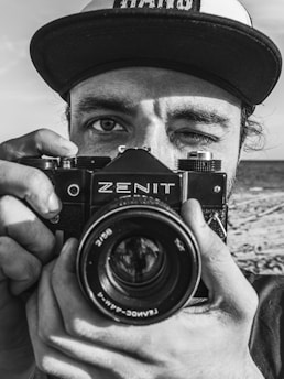 A person wearing a cap is holding a vintage Zenit camera in front of their face, focusing the lens directly at the viewer. The person's right eye is closed, suggesting they are in the act of taking a photograph. The background appears to be a beach with sand visible.