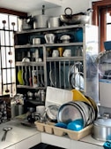 A kitchen setup featuring a metal rack holding an assortment of stainless steel pots, pans, and plates. There are various kitchen utensils and containers arranged on a counter below. The setting appears organized yet busy, with a mix of metal, plastic, and ceramic items.