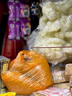 A market stall features a variety of packaged snacks. There is a large orange plastic bag filled with crunchy noodles. Nearby, a transparent mesh bag contains lighter-colored puff snacks. Additionally, a stack of rectangular packaged snacks is visible, along with hanging colorful snack packets in the background.