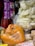 A market stall features a variety of packaged snacks. There is a large orange plastic bag filled with crunchy noodles. Nearby, a transparent mesh bag contains lighter-colored puff snacks. Additionally, a stack of rectangular packaged snacks is visible, along with hanging colorful snack packets in the background.