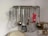 On a wall-mounted rack, various kitchen utensils are hanging, including a whisk, ladle, grater, spatula, and strainer. The background has a noticeable texture and discoloration. A mix of metallic and red plastic elements is visible, with an outlet and some wiring in the corner.