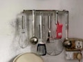On a wall-mounted rack, various kitchen utensils are hanging, including a whisk, ladle, grater, spatula, and strainer. The background has a noticeable texture and discoloration. A mix of metallic and red plastic elements is visible, with an outlet and some wiring in the corner.