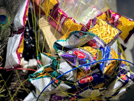 A variety of snack packets, primarily Haldiram's brand, are tied together with netting. The packets come in different colors, mostly yellow, purple, and green, and are wrapped in a circular pattern. A peacock feather adds a decorative element on the side.