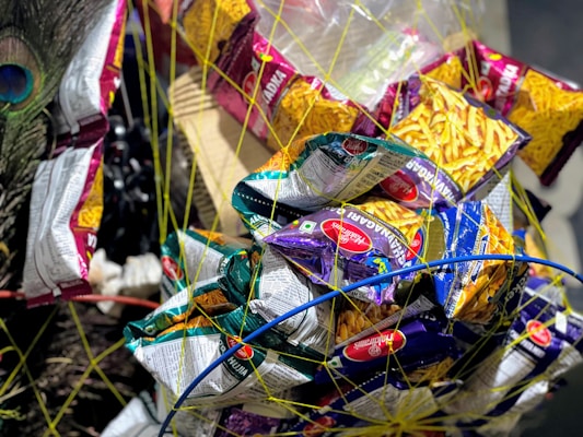 A variety of snack packets, primarily Haldiram's brand, are tied together with netting. The packets come in different colors, mostly yellow, purple, and green, and are wrapped in a circular pattern. A peacock feather adds a decorative element on the side.