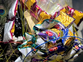 A variety of snack packets, primarily Haldiram's brand, are tied together with netting. The packets come in different colors, mostly yellow, purple, and green, and are wrapped in a circular pattern. A peacock feather adds a decorative element on the side.
