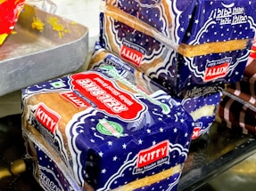 Packages of sliced bread with a blue and white design marked with the label 'Kitty Real Bake' are placed on a surface. The packaging features stars and has prominent red and green logos, alongside a 'KITTY The Master Baker' label. Part of a metal tray with residual yellow substance is visible to the side.