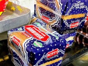Packages of sliced bread with a blue and white design marked with the label 'Kitty Real Bake' are placed on a surface. The packaging features stars and has prominent red and green logos, alongside a 'KITTY The Master Baker' label. Part of a metal tray with residual yellow substance is visible to the side.