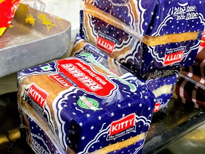 Packages of sliced bread with a blue and white design marked with the label 'Kitty Real Bake' are placed on a surface. The packaging features stars and has prominent red and green logos, alongside a 'KITTY The Master Baker' label. Part of a metal tray with residual yellow substance is visible to the side.