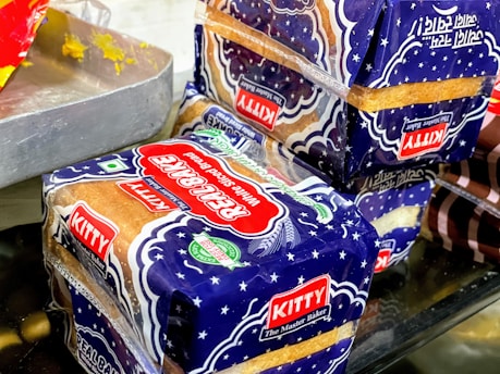 Packages of sliced bread with a blue and white design marked with the label 'Kitty Real Bake' are placed on a surface. The packaging features stars and has prominent red and green logos, alongside a 'KITTY The Master Baker' label. Part of a metal tray with residual yellow substance is visible to the side.