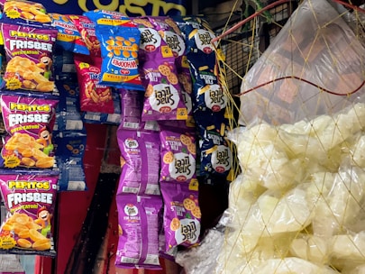 Various snack packages hang on a display, featuring colorful designs. The packages include brands like Pepitos Frisbee and Fun Wheels, with vibrant purple, blue, orange, and red colors. On the right, clear plastic bags contain white snack items, possibly popcorn or puffed chips, held together by a net.