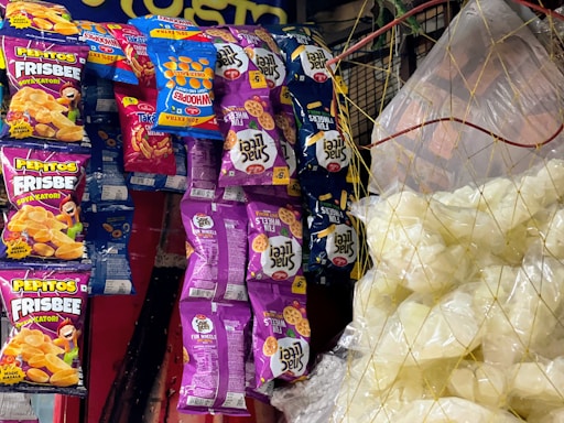 Various snack packages hang on a display, featuring colorful designs. The packages include brands like Pepitos Frisbee and Fun Wheels, with vibrant purple, blue, orange, and red colors. On the right, clear plastic bags contain white snack items, possibly popcorn or puffed chips, held together by a net.