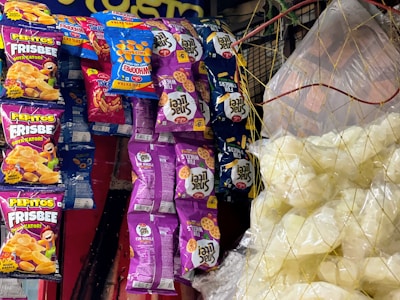 Various snack packages hang on a display, featuring colorful designs. The packages include brands like Pepitos Frisbee and Fun Wheels, with vibrant purple, blue, orange, and red colors. On the right, clear plastic bags contain white snack items, possibly popcorn or puffed chips, held together by a net.