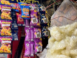 Various snack packages hang on a display, featuring colorful designs. The packages include brands like Pepitos Frisbee and Fun Wheels, with vibrant purple, blue, orange, and red colors. On the right, clear plastic bags contain white snack items, possibly popcorn or puffed chips, held together by a net.