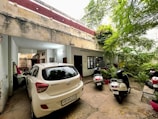 A partially covered parking area is visible, with a white car in the foreground and two scooters parked nearby. The area is bordered by an old building with peeling paint and a staircase leading to an upper floor. Lush greenery, including trees and plants, is visible on the right side, contributing to a natural and earthy atmosphere.