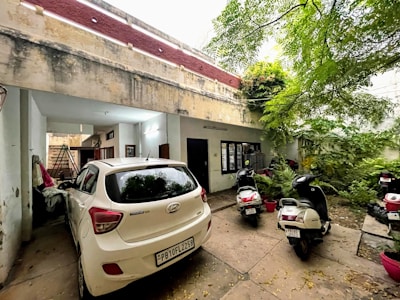 A partially covered parking area is visible, with a white car in the foreground and two scooters parked nearby. The area is bordered by an old building with peeling paint and a staircase leading to an upper floor. Lush greenery, including trees and plants, is visible on the right side, contributing to a natural and earthy atmosphere.