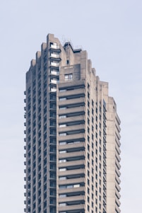 A tall, concrete skyscraper with a brutalist architectural style, featuring numerous balconies on each floor, and windows symmetrically arranged around the structure. The building’s surface is composed of exposed concrete, giving it a rugged and industrial appearance. The sky in the background is clear and pale blue.