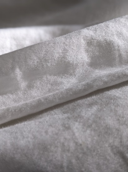 A close-up view of a piece of fabric with a soft texture, displaying a gentle sheen. The material appears to be folded, forming subtle shadows and highlights on its surface.