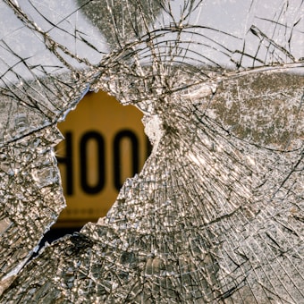 A broken glass with a large central crack and web-like fractures radiating outward. Behind the shattered glass, there is a blurred object with the word 'HOOD' partially visible in bold letters.
