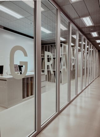 A modern office interior with a series of glass doors. Large letters are printed on the doors, possibly forming a word or acronym. Inside, there are shelves filled with organized materials, possibly papers or files. The ceiling has built-in lights providing a bright, well-lit environment.