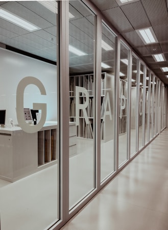 A modern office interior with a series of glass doors. Large letters are printed on the doors, possibly forming a word or acronym. Inside, there are shelves filled with organized materials, possibly papers or files. The ceiling has built-in lights providing a bright, well-lit environment.