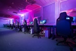 A row of gaming computers with large monitors and racing-style chairs is set up along the interior of a modern room. The space is illuminated with vibrant purple and blue LED lighting, creating a futuristic atmosphere. The room is organized and clean, suggesting a professional gaming or esports environment.
