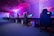 A row of gaming computers with large monitors and racing-style chairs is set up along the interior of a modern room. The space is illuminated with vibrant purple and blue LED lighting, creating a futuristic atmosphere. The room is organized and clean, suggesting a professional gaming or esports environment.