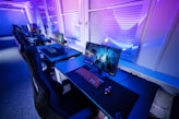 A row of modern gaming setups is arranged neatly along a window-lined wall. Each station includes a high-resolution monitor displaying vivid graphics, a mechanical keyboard with colored lighting, a gaming mouse, and an ergonomic chair. The room is illuminated with ambient blue and purple lights, creating a sleek and futuristic atmosphere.
